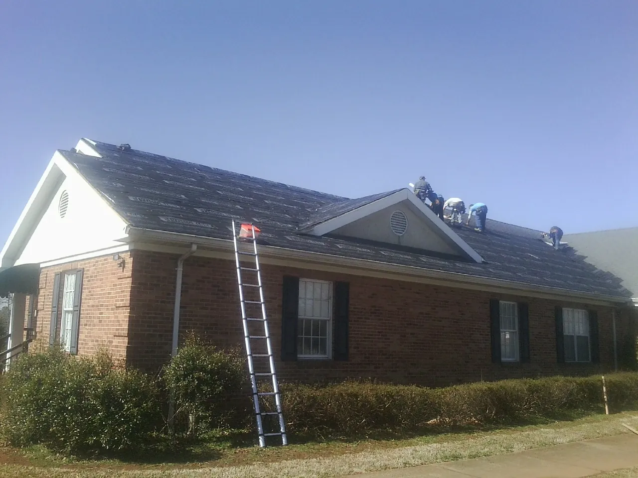 Crew tearing shingles off a brick ranch roof with a ladder set — Metal Roof Repair work by our crew in Almont